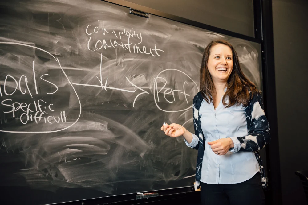 UCONN MBA instructor standing in front of chalkboard smiling teaching course.