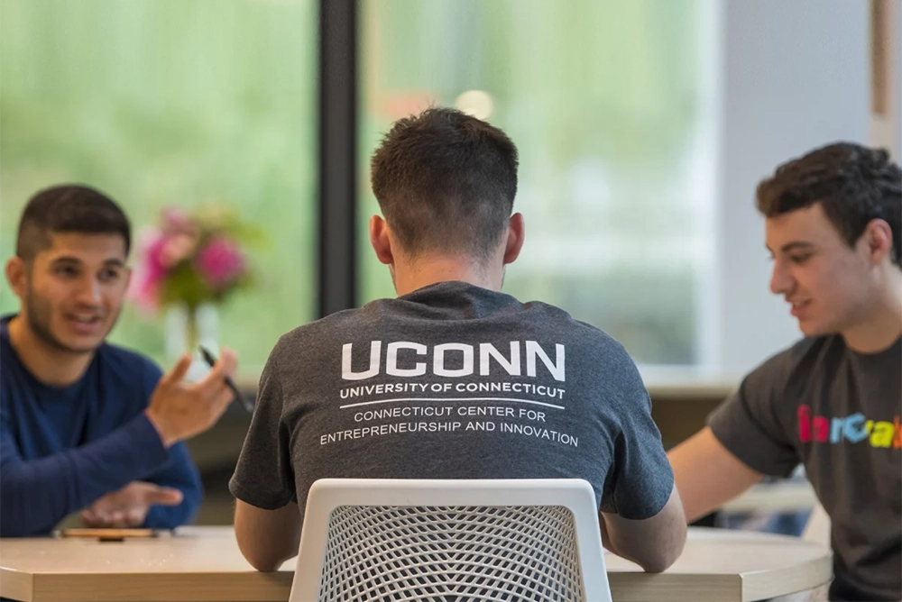 Three college students seated at a table in conversation with focus on the back of one student showing a tshirt with the UConn Connecticut Center for Entrepreneurship and Innovation wordmark