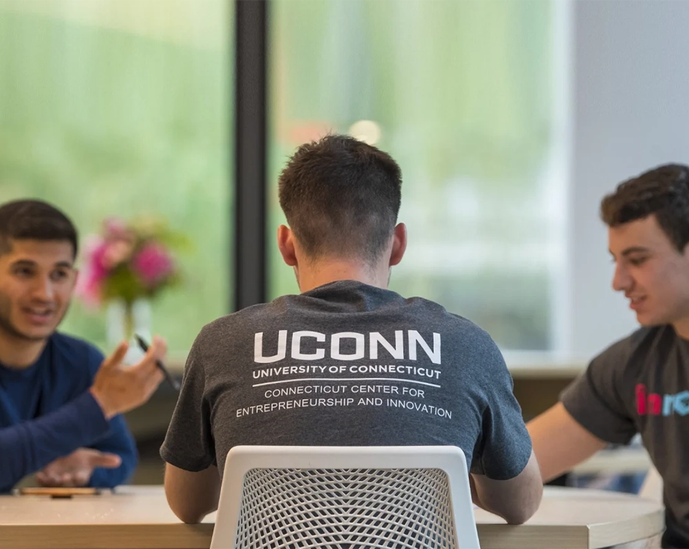 Three college students seated at a table in conversation with focus on the back of one student showing a tshirt with the UConn Connecticut Center for Entrepreneurship and Innovation wordmark