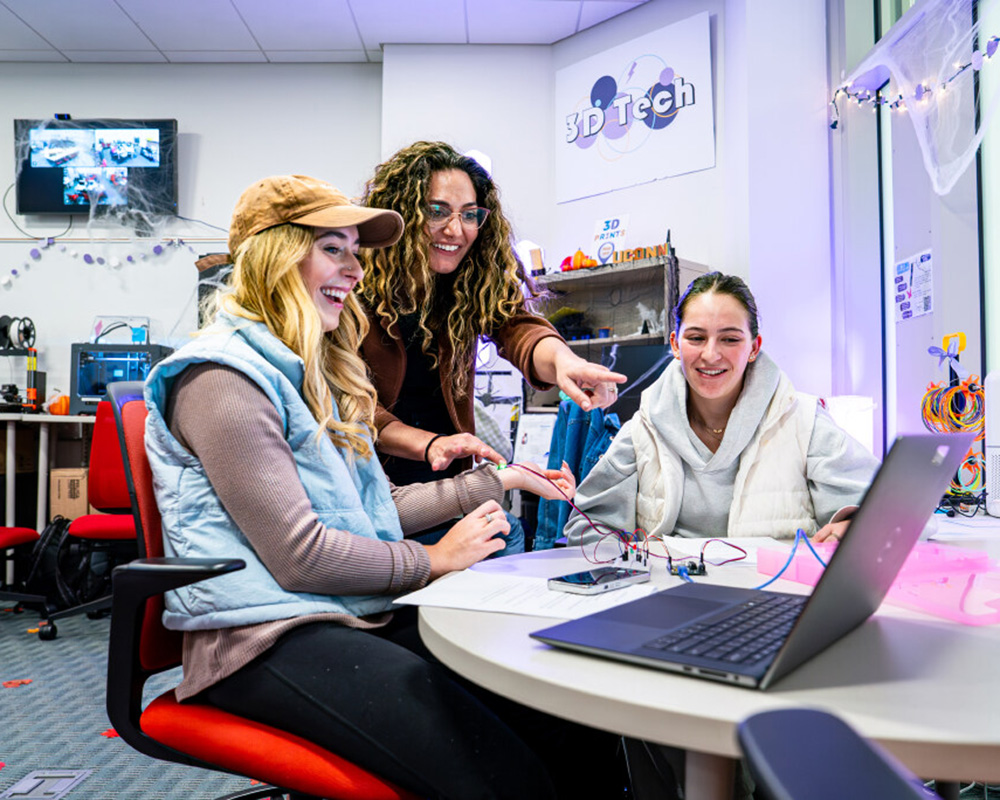 Three students in a School of Business technology lab gathered around a laptop pointing at the screen with enthusiasm