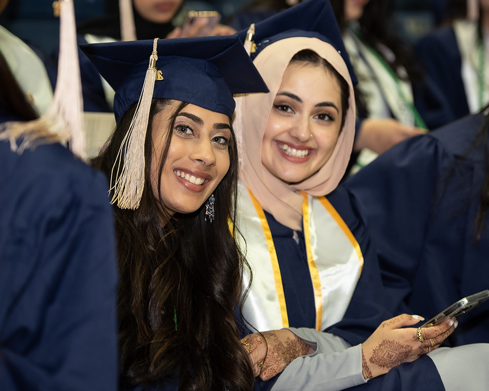 Two smiling business students seated among the crowd at a graduation ceremony